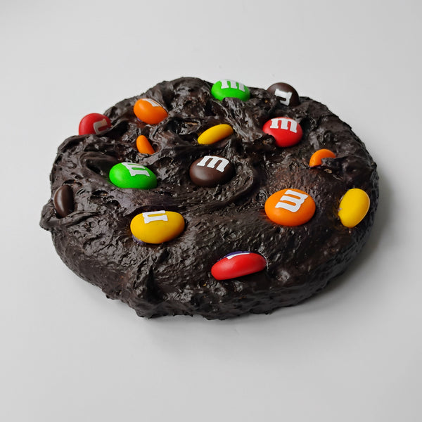A chocolate chip cookie decorated with colorful M&M's candies, photographed on a neutral light gray background.