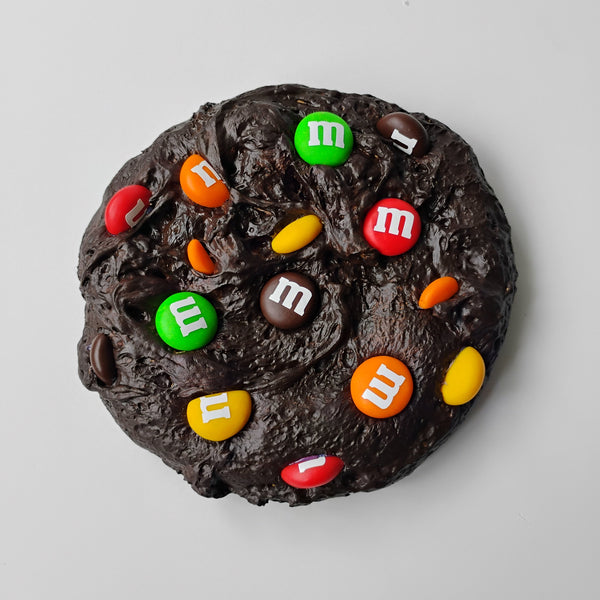 A chocolate brownie cookie topped with colorful M&M's candies, photographed from directly above on a light gray wall