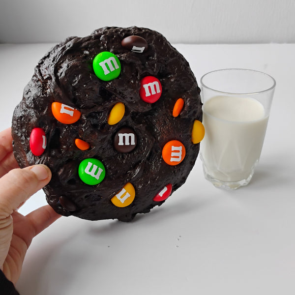 A hand holds a giant chocolate brownie decorated with colorful M&M's candies, next to a glass of milk on a white surface.