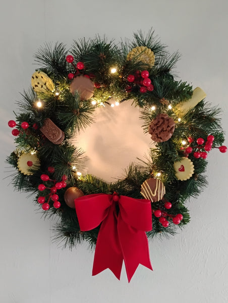 Decorative Christmas wreath with lights, NZ biscuits, berries, and a red bow on a plain background.